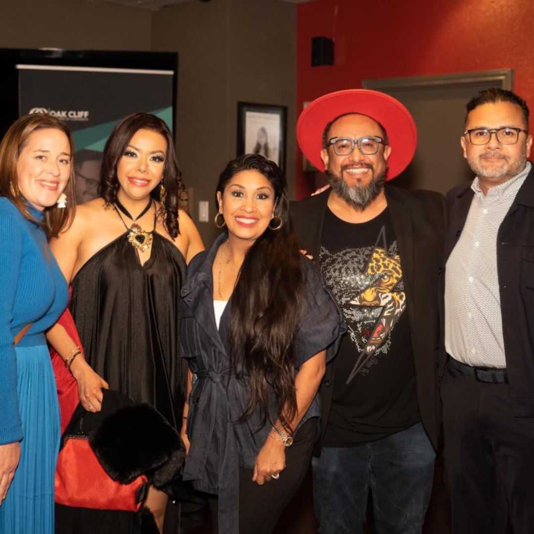 Mexican cultural shop team that make a mark in this world. The five people are in a conference room and behind it is an orange wall and a digital sign. They are dressed in elegant black and blue outfits with accessories such as hats and handmade necklaces