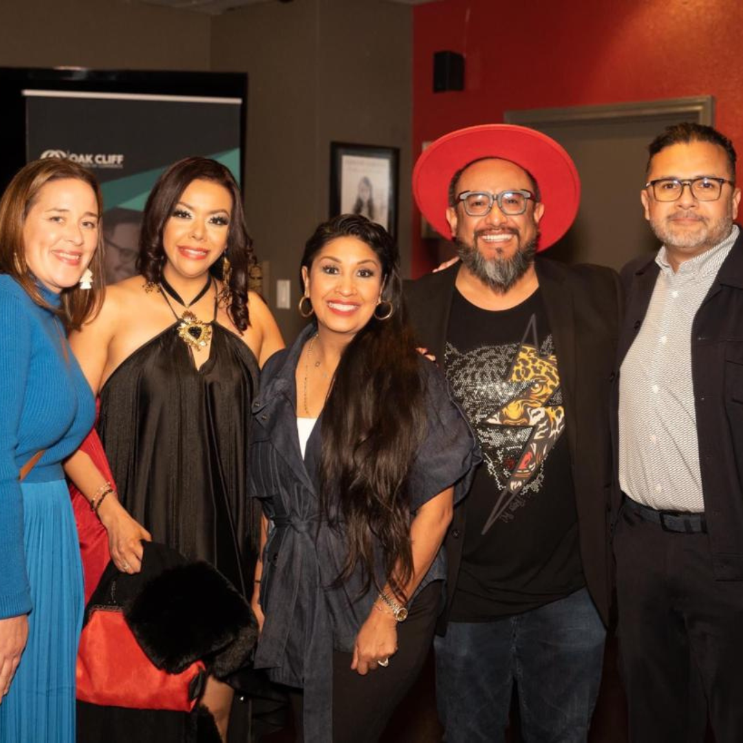 Mexican cultural shop team that make a mark in this world.  The five people are in a conference room and behind it is an orange wall and a digital sign. They are dressed in elegant black and blue outfits with accessories such as hats and handmade necklaces