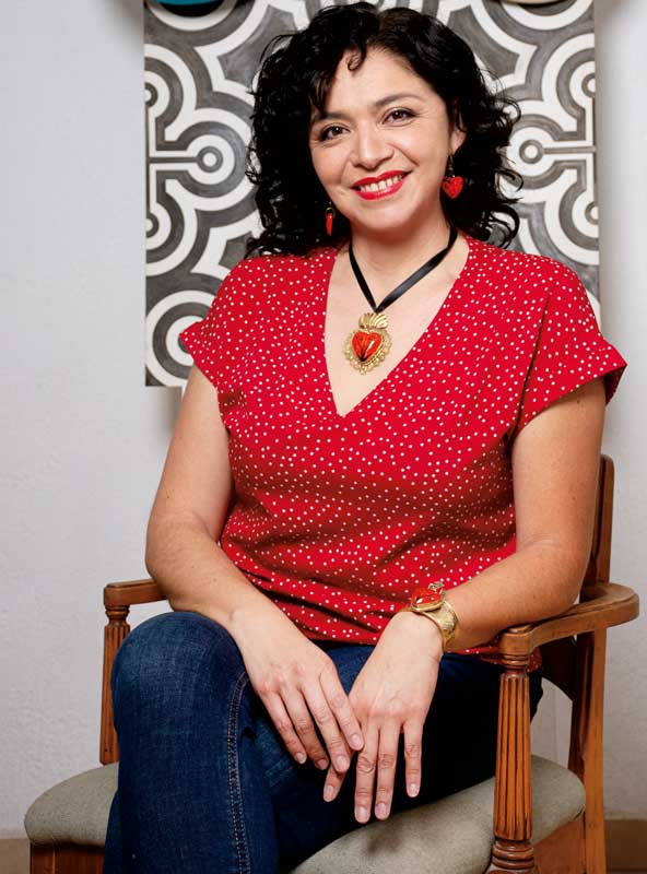 The handcrafting artisan is sitting on a wooden chair, in front of a white wall and an abstract art painting.  She is dressed in a red blouse with small white dots and dark blue jeans. As accessories she has a handmade red heart-shaped necklace and earrings