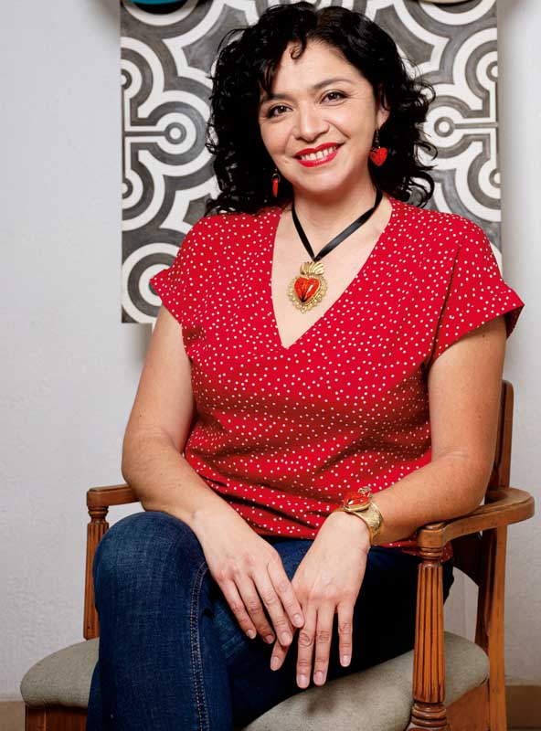 The handcrafting artisan is sitting on a wooden chair, in front of a white wall and an abstract art painting. She is dressed in a red blouse with small white dots and dark blue jeans. As accessories she has a handmade red heart-shaped necklace and earrings