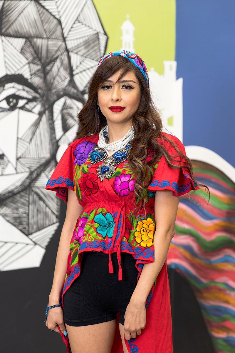 World fashion long-tailed red blouse with purple, pink and yellow fabric in flowers and leaves. The woman is wearing multicultural accessories and behind her is a wall with colorful illustrations