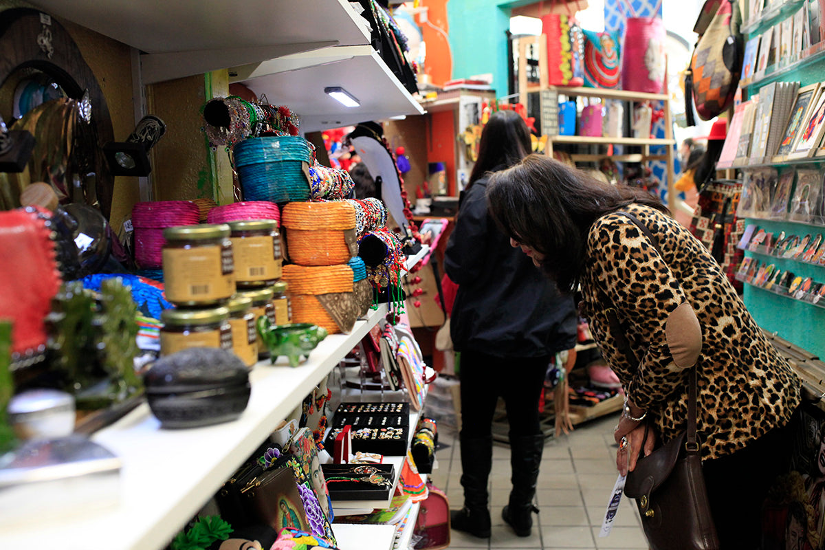In the multicultural store there are two women buying handmade products. On the shelves are bags embroidered with the image of Frida Kahlo, accessories such as bracelets, necklaces and rings, orange and blue woven baskets
