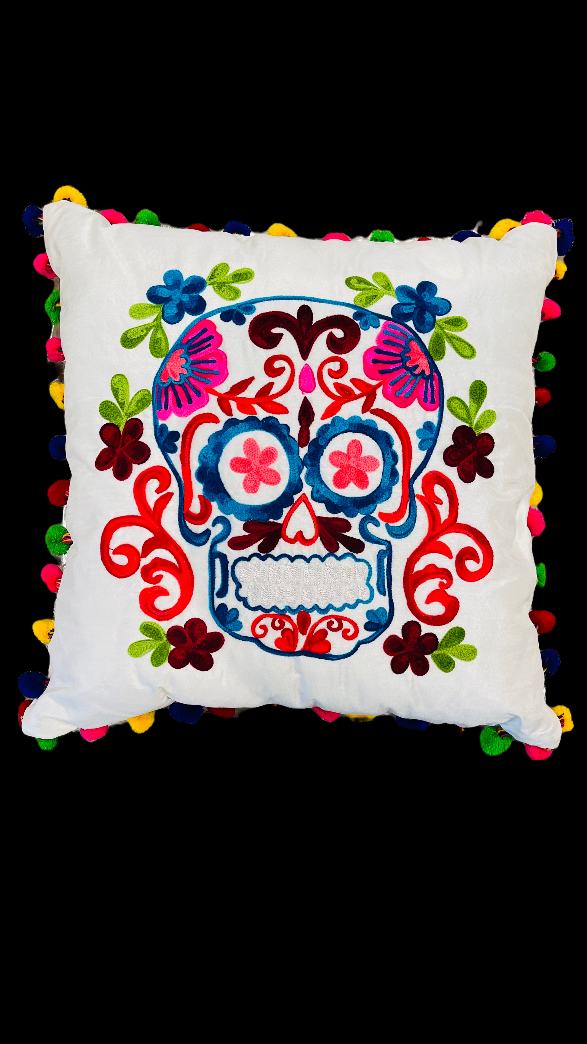 Hand embroidered white pillow with a skull in the center in pink, orange, blue and white with green leaves around it. The border of the cushion is made with pompoms of different colors.