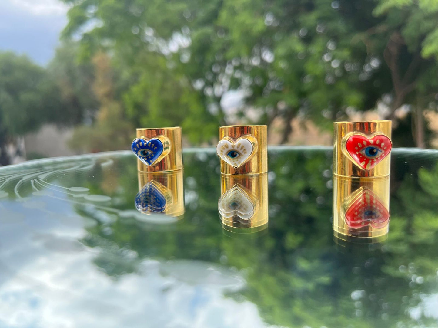 Hand-painted cultural fashion rings in the shape of a red, blue and white heart. In the middle are painted golden eyes and they are on a glass dining room with green trees behind.