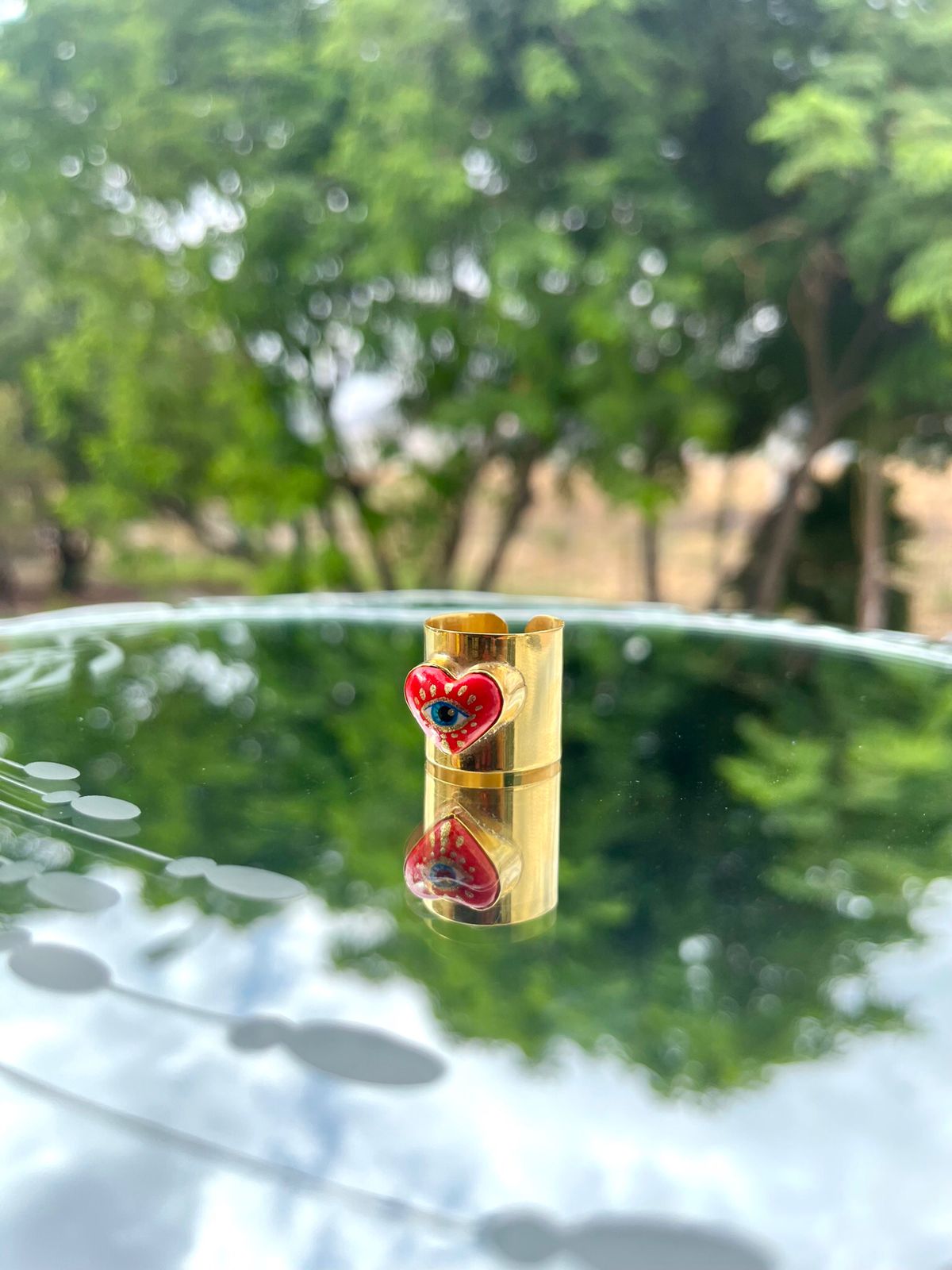 Hand-painted cultural fashion gold ring in the shape of a red heart. In the center is painted an eye, and it is on a round glass dining room.