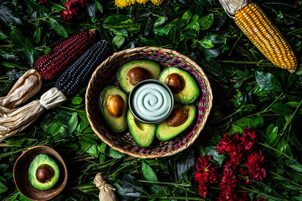 The avocado styling cream is inside a handmade bowl with five avocados underneath. Around the cream there are green leaves and pink flowers and petals all around