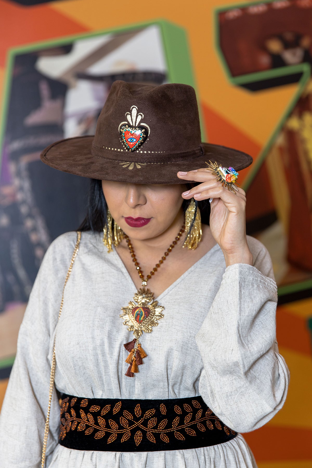 A woman in a handmade white blouse is wearing a brown hat inspired by Mexican culture. In the center of the hat is a famous red carazon with a painted eye, and she is accessorized with a gold virgin of guadalupe necklace and long earrings. On the right hand she has a white catrina ring made with paper mache