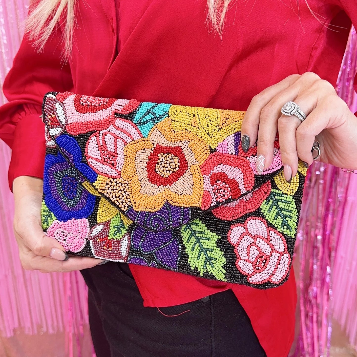 The colorful beaded bag is made with intricate black, red, pink, green and blue stones. It is designed with flowers and is held by a woman in a red shirt with a beautiful ring in her hand.