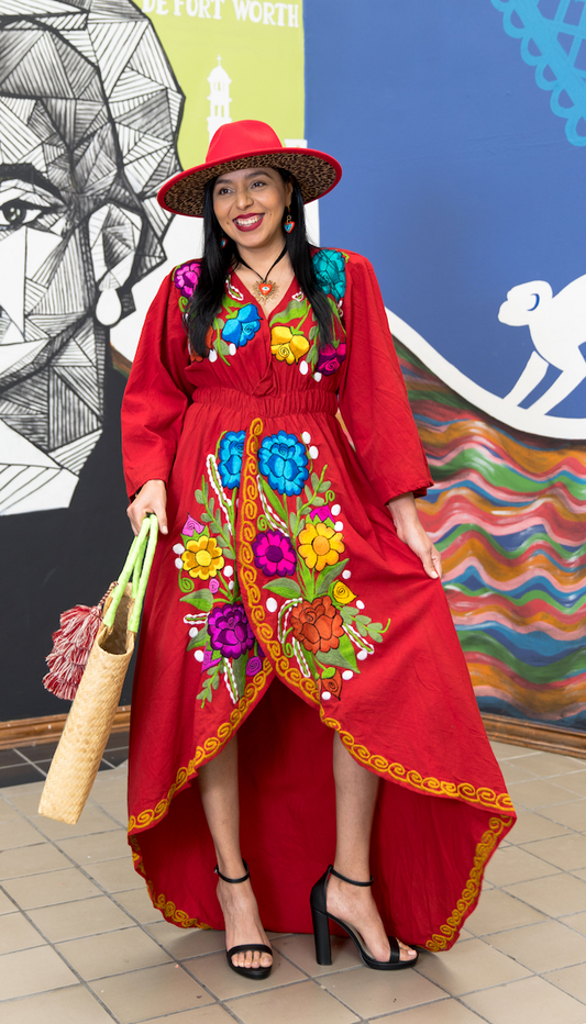 Woman dressed in long-tailed red dress with embroidered flowers of different sizes and colors. On her head she has a red hat and a handmade bag in her left hand. 