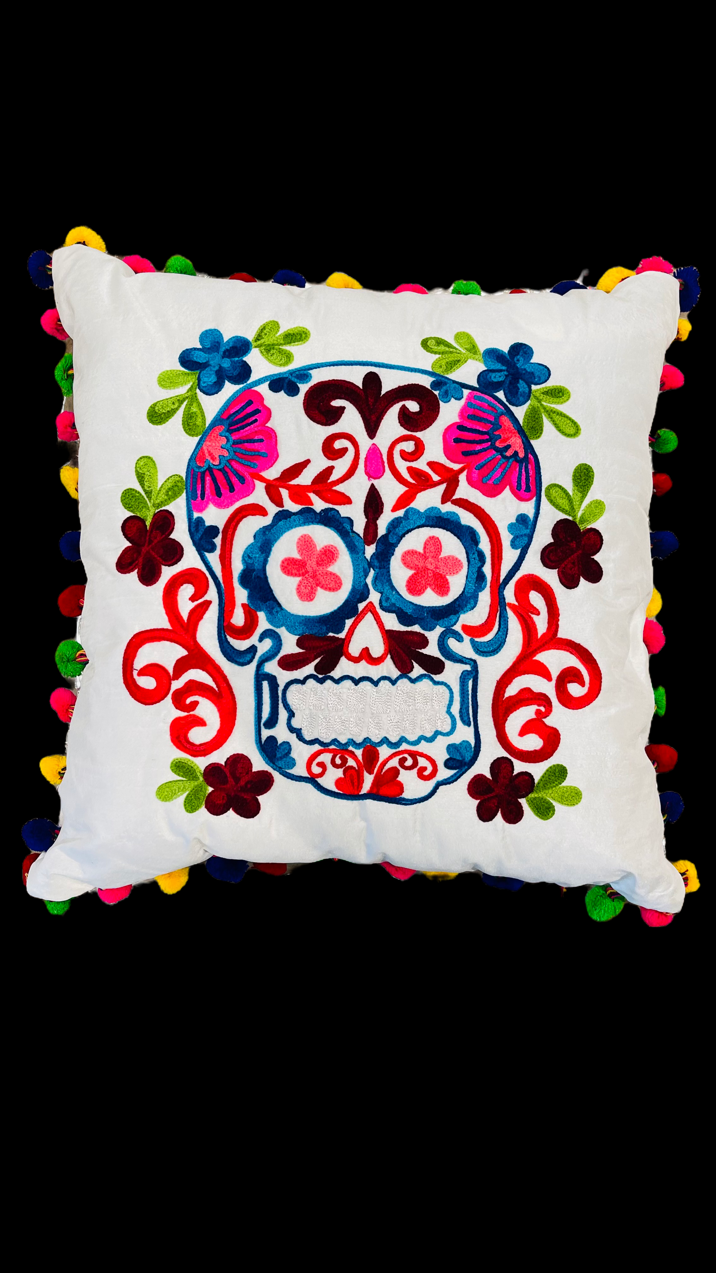 Hand embroidered white pillow with a skull in the center in pink, orange, blue and white with green leaves around it. The border of the cushion is made with pompoms of different colors. 