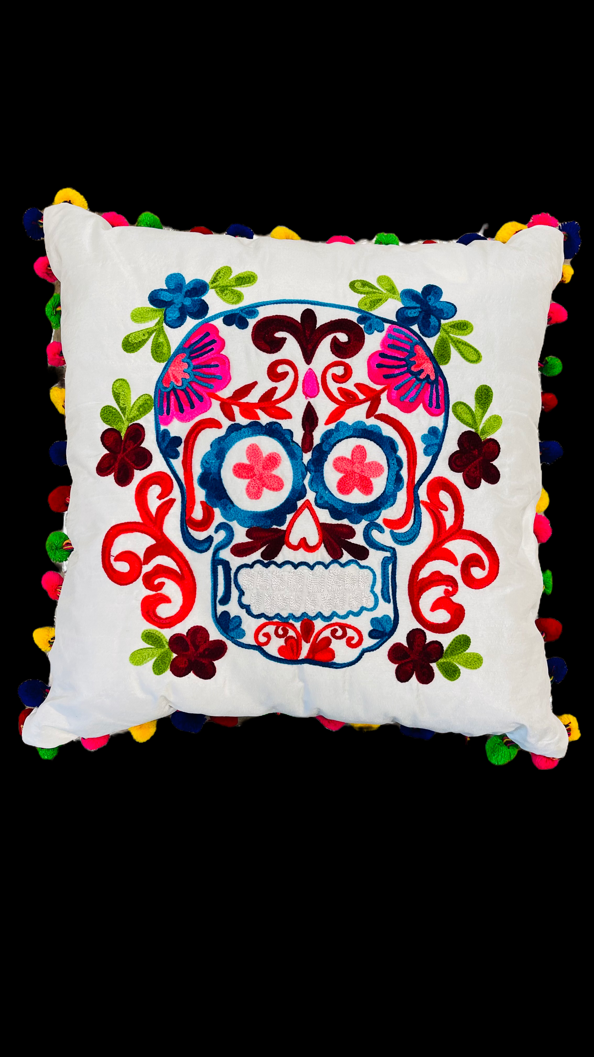 Hand embroidered white pillow with a skull in the center in pink, orange, blue and white with green leaves around it. The border of the cushion is made with pompoms of different colors. 