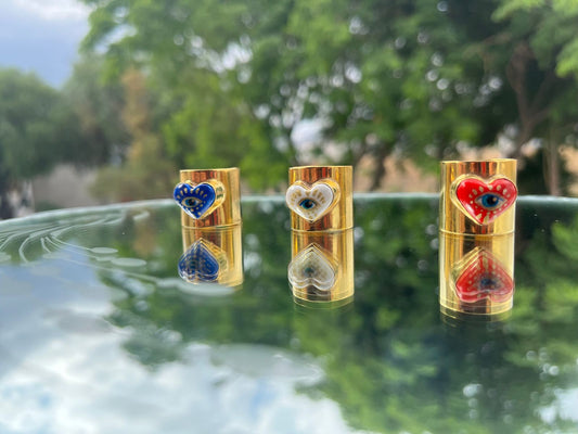 Hand-painted cultural fashion rings in the shape of a red, blue and white heart. In the middle are painted golden eyes and they are on a glass dining room with green trees behind.