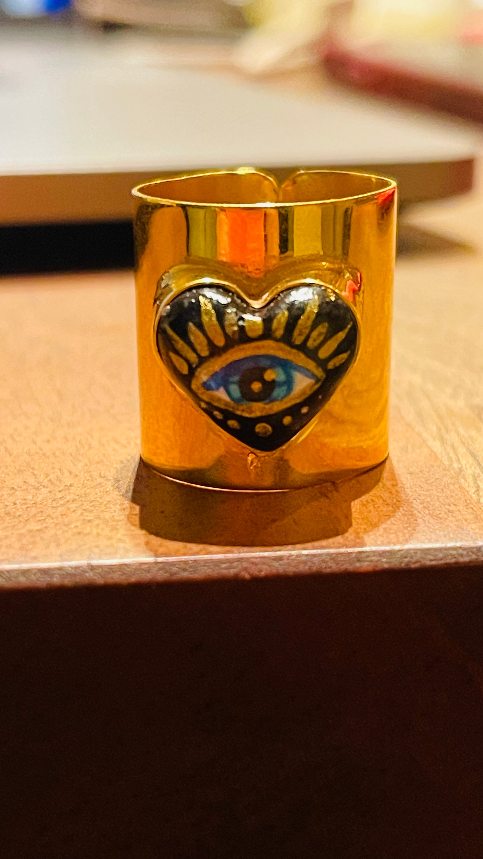 Hand-painted cultural fashion gold ring in the shape of a black heart. In the middle is painted an eye, and it is on a wooden table.