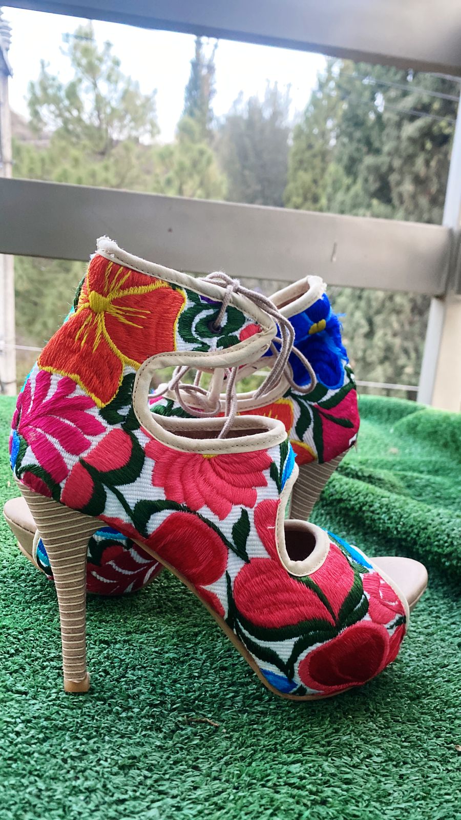 High heels with blue, red and yellow floral embroidery. The heels are on grass and there is a balcony behind them.